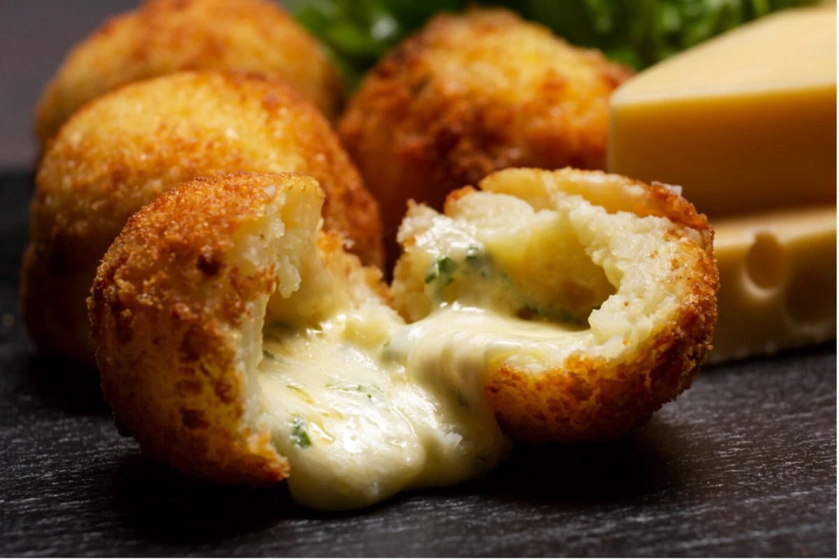 Zbliżenie na przekrojoną na pół kulkę arancini z wylewającym się serem, w tle widać kulki w całości